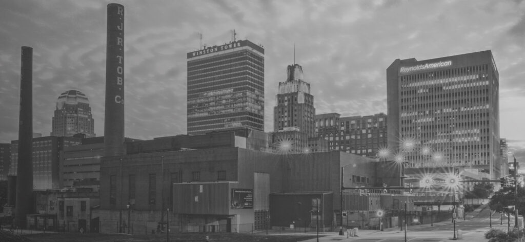 From the left to the right of this skyline photograph of Winston-Salem, there are two brick smokestacks and several skyscrapers that make up the downtown area.