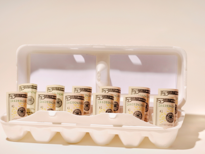 On a pale pink background, is a carton of eggs filled with rolled up five dollar bills.