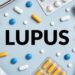 The word Lupus is surrounded by blue, orange and white pills, pill packages, a stethoscope and a bandage wrap.
