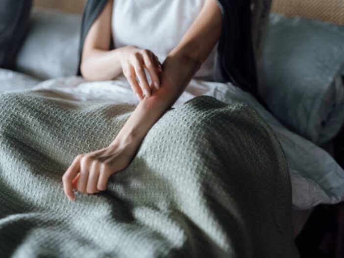A person sitting up in bed scratches their left forearm.