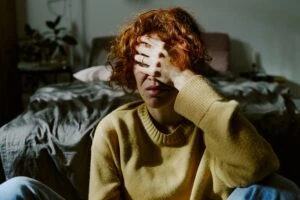 Woman sitting in a dark bedroom with her face in her hands. This is the header image for the blog post titled - "Can Anxiety or Depression Qualify for Disability?"