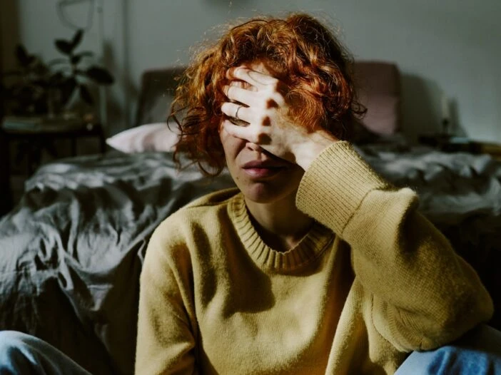 Woman sitting in a dark bedroom with her face in her hands. This is the header image for the blog post titled - "Can Anxiety or Depression Qualify for Disability?"