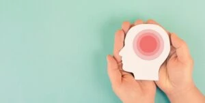 The image has two human hands holding an object that looks like a human head with a red circle in the center which symbolizes headache pain. This image accompanies a blog post titled - Can You Get Disability for Chronic Migraines in Charlotte, NC?