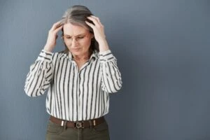 Picture of an older woman with grey hair and glasses holding her head. She's standing against a grey background and the picture is associated with a blog post titled - "Can You Get Disability for Sjögren’s Syndrome in Charlotte, NC?"