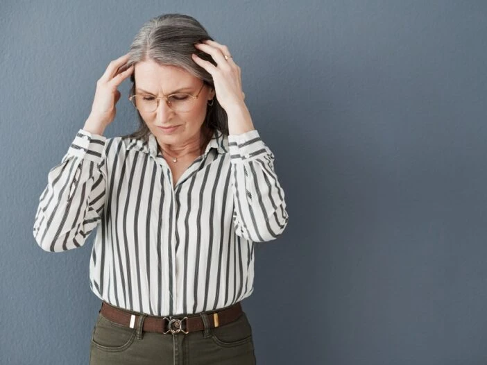Picture of an older woman with grey hair and glasses holding her head. She's standing against a grey background and the picture is associated with a blog post titled - "Can You Get Disability for Sjögren’s Syndrome in Charlotte, NC?"