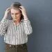 Picture of an older woman with grey hair and glasses holding her head. She's standing against a grey background and the picture is associated with a blog post titled - "Can You Get Disability for Sjögren’s Syndrome in Charlotte, NC?"