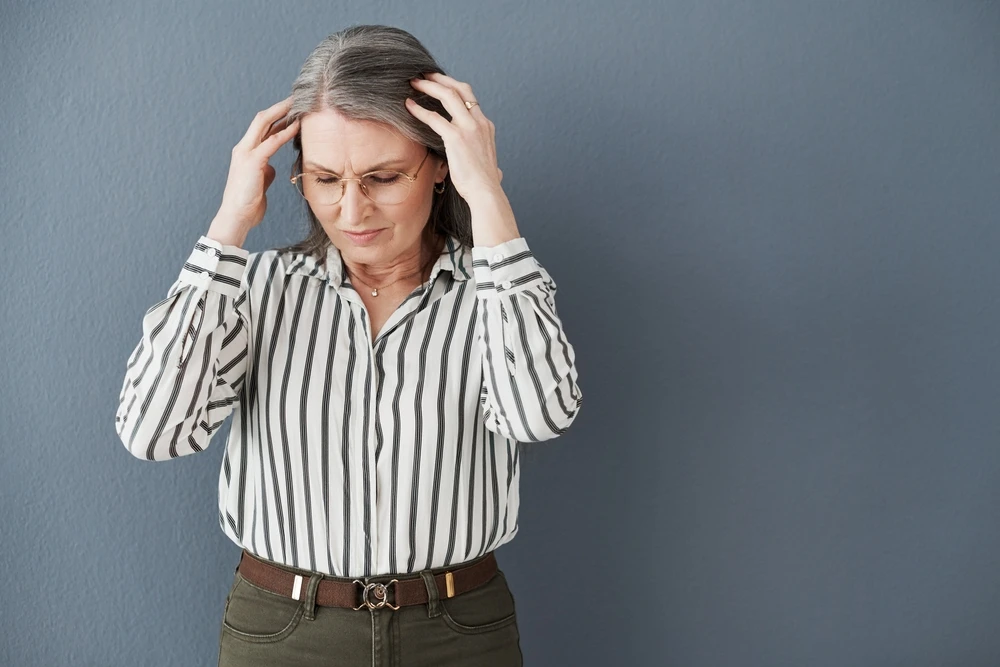 Picture of an older woman with grey hair and glasses holding her head. She's standing against a grey background and the picture is associated with a blog post titled - "Can You Get Disability for Sjögren’s Syndrome in Charlotte, NC?"
